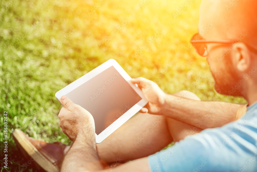 Close-up of casual dressed young man using modern digital tablet while sitting at the park, sun light