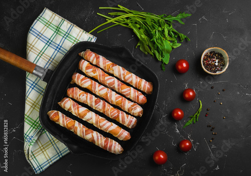 Preparation of raw sausages wrapped spirally in bacon on a cast-iron frying pan. Ingredients for sausages in bacon cherry tomatoes, rucola salad. Black concrete background. Top view, flat lay.