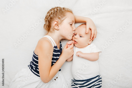 Lifestyle portrait of cute white Caucasian girl sister holding kissing little baby, lying on bed indoors. Older sibling with younger brother newborn. Family love bonding together concept.