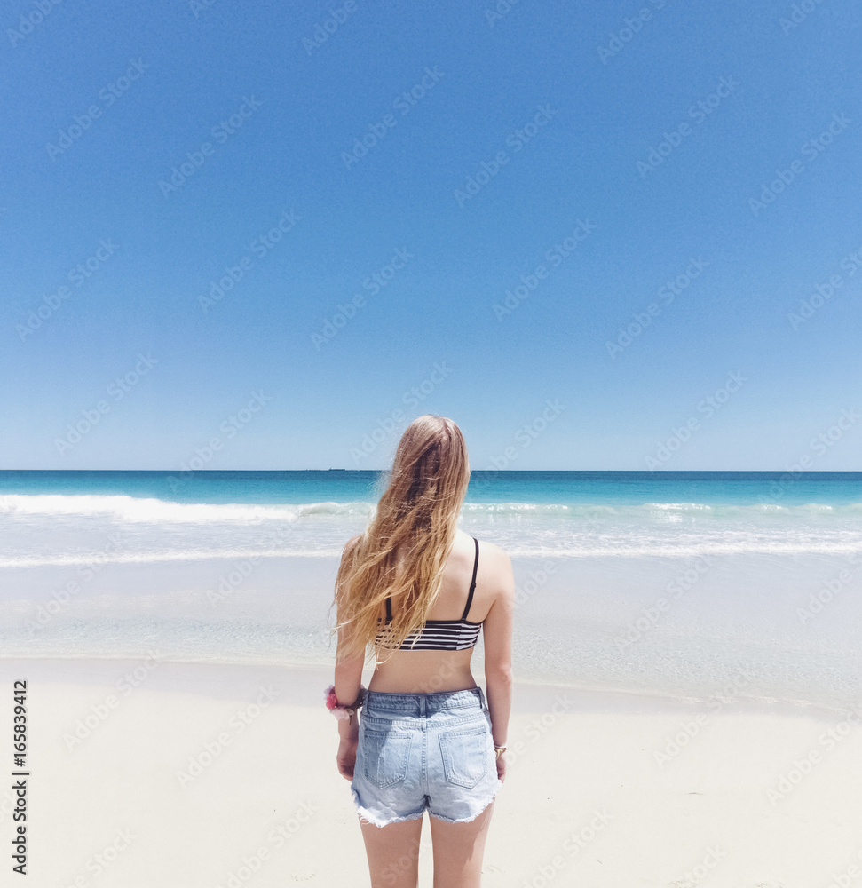 Back view of young woman, with long blonde hair, facing the ocean Back view of young woman, with long blonde hair, facing the ocean