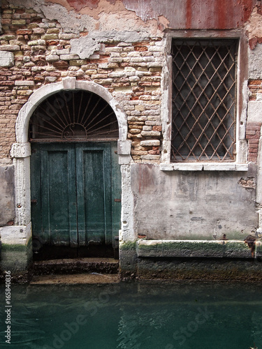 old green door in an ancient brick building with window and arched doorway on a canal in venice with reflection in the water damp walls and crumbling painted plaster © philopenshaw