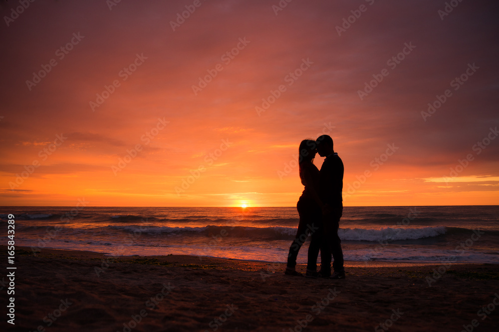 Couple in love having romantic tender moments at sunset. Honeymoon lovers hugging on beach. Silhouettes of couple against the sunset sky.