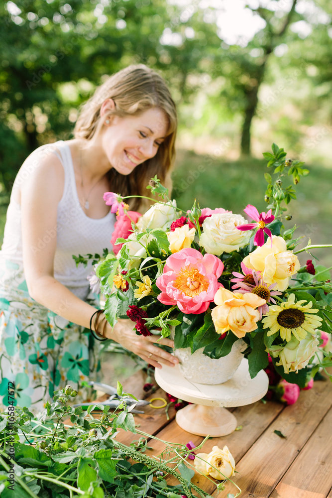 bouquet, people and floral arrangement concept - smiling young woman florist with bouquet of beautiful peonies, daisies, yellow and white roses in white vase at the wooden table in summer garden bouquet, people and floral arrangement concept - smiling young woman florist with bouquet of beautiful peonies, daisies, yellow and white roses in white vase at the wooden table in summer garden