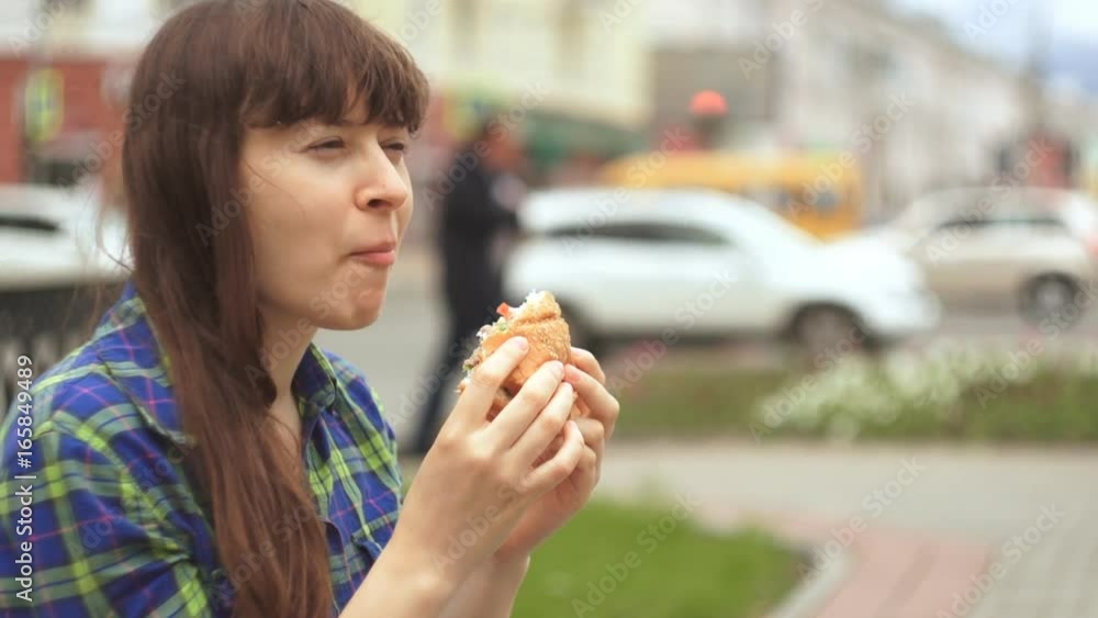 Close-up shot of a young attractive brunette woman in a plaid shirt eating a hamburger outdoors on a background of brisk traffic of people on the road and cars on the freeway. Close-up shot of a young attractive brunette woman in a plaid shirt eating a hamburger outdoors on a background of brisk traffic of people on the road and cars on the freeway.