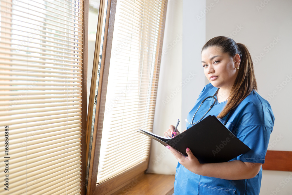 Young female hispanic doctor reading and filling a report form, standing next to a window in a hospital hallway Young female hispanic doctor reading and filling a report form, standing next to a window in a hospital hallway