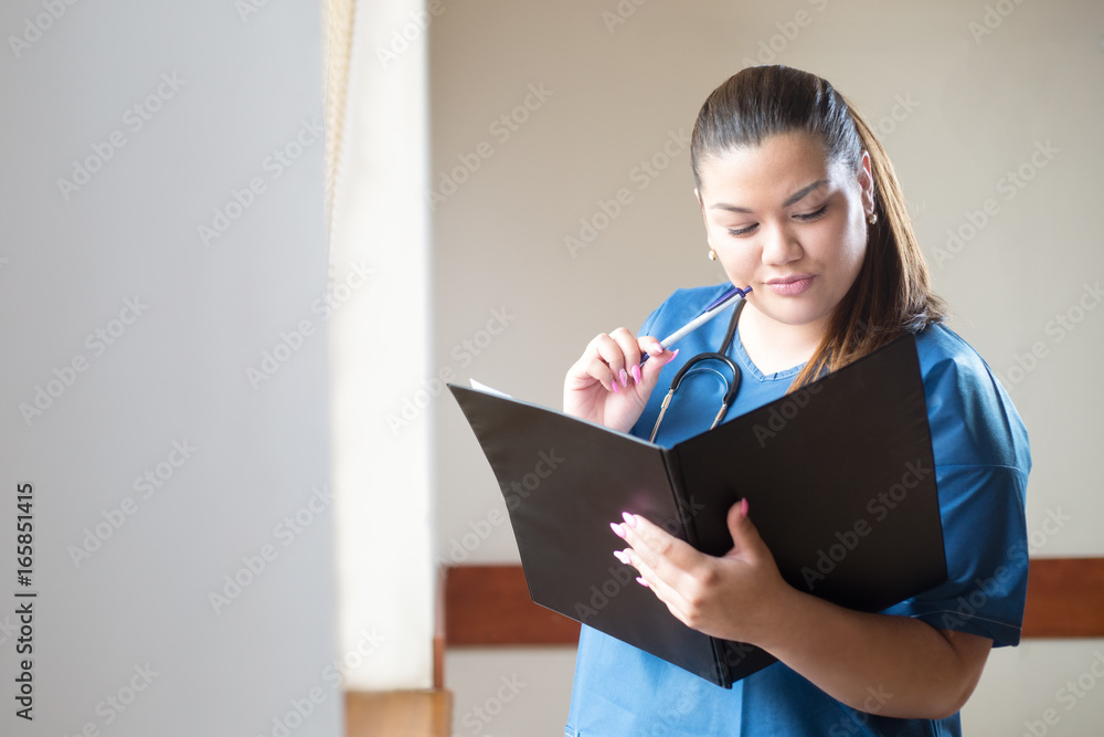Young nurse frown while analyzing a clinical case, looking into a folder she is holding in her hands. Woman is wearing a blue uniform, standing in hospital next to a window. Young nurse frown while analyzing a clinical case, looking into a folder she is holding in her hands. Woman is wearing a blue uniform, standing in hospital next to a window.