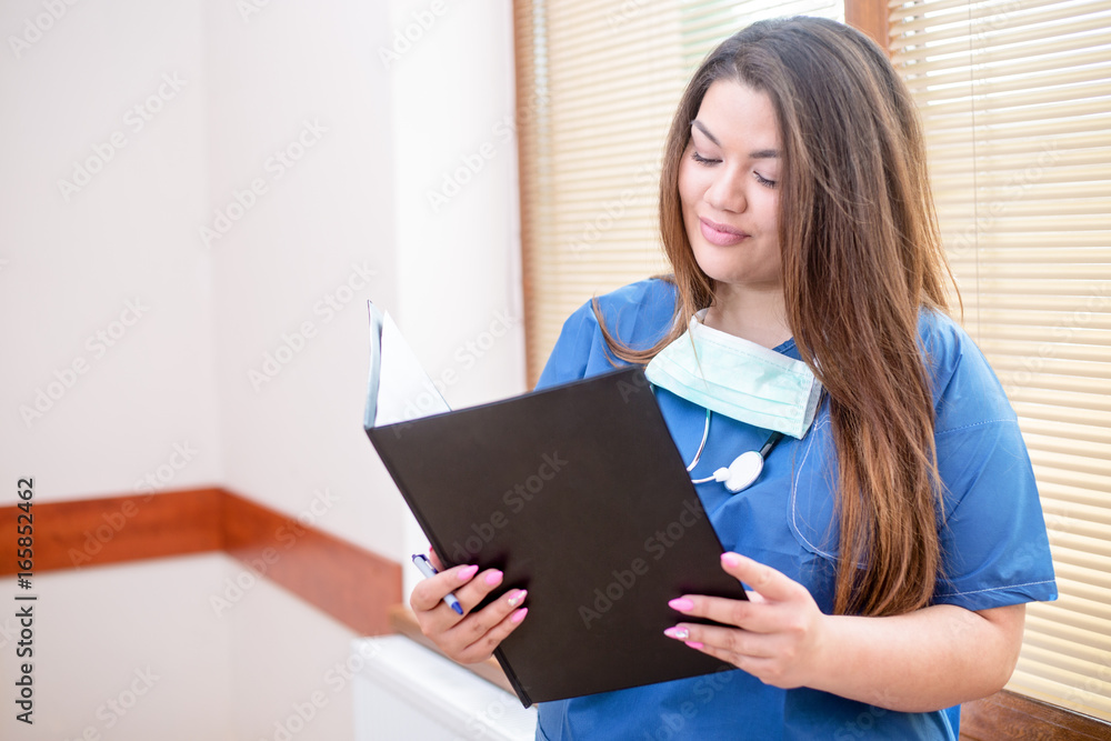 Smiling confident medical female doctor standing in a hospital hallway next to a window, wearing a blue uniform and stethoscope, looking into her folder Smiling confident medical female doctor standing in a hospital hallway next to a window, wearing a blue uniform and stethoscope, looking into her folder