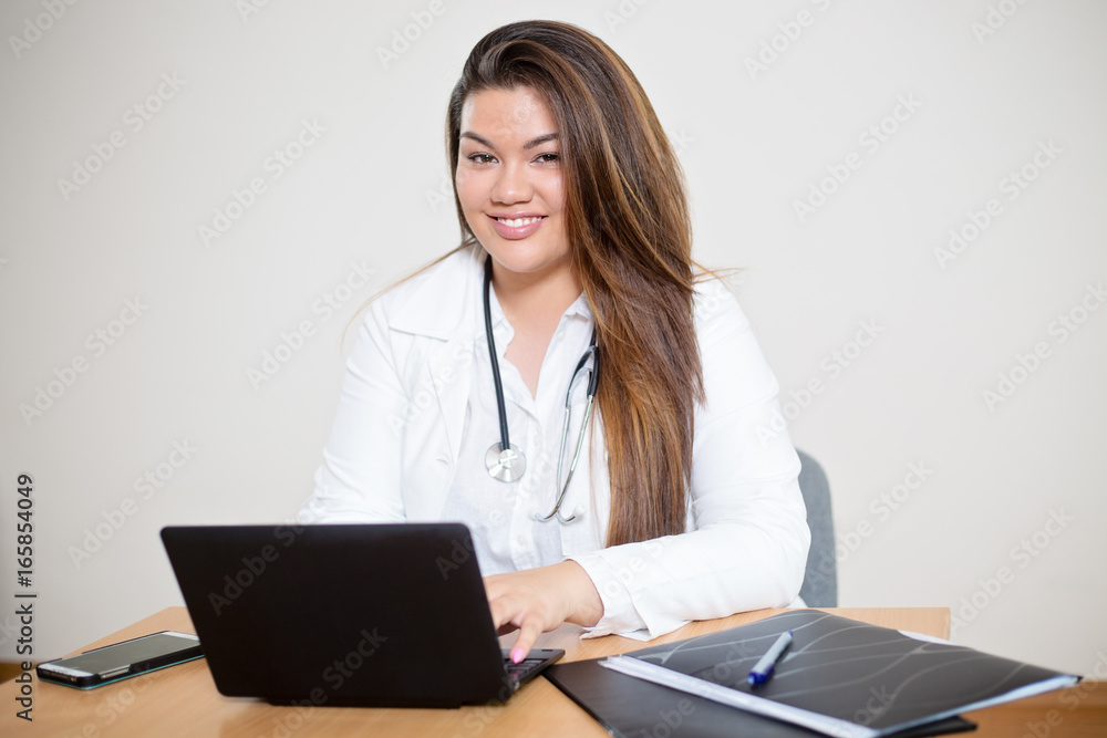 Young female hispanic doctor sitting behind the desk, typing into a laptop, looking into a camera Young female hispanic doctor sitting behind the desk, typing into a laptop, looking into a camera
