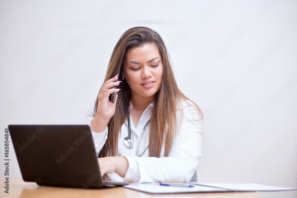 Young female hispanic doctor sitting behind the desk, typing into a laptop, talking on the mobile phone and looking into a paper documents on her table Young female hispanic doctor sitting behind the desk, typing into a laptop, talking on the mobile phone and looking into a paper documents on her table