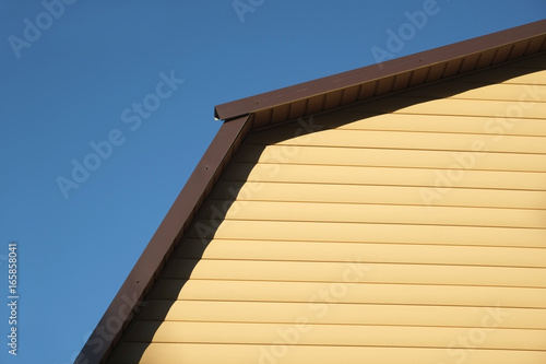Part of rural house Wall covered with yellow siding and brown metal roof on clear blue cloudless sky on sunny day front view closeup