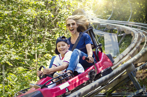 Photography Smiling women and her boy riding downhill together on an outdoor roller coaster on a warm summer day