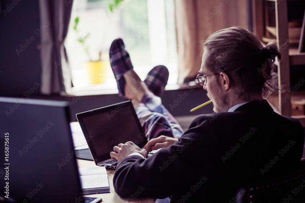 Rear view of freelancer at working process. Young man working at wooden table, his legs in pajamas trousers and slippers. Toned image. - obrazy, fototapety, plakaty 