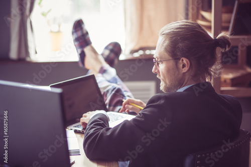 Man working at home office. Freelancer working with computer and laptop with his feet in slippers on wooden table. Toned image.