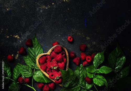 Bowl iwith fresh raspberries and green leaves on a dark background, top view