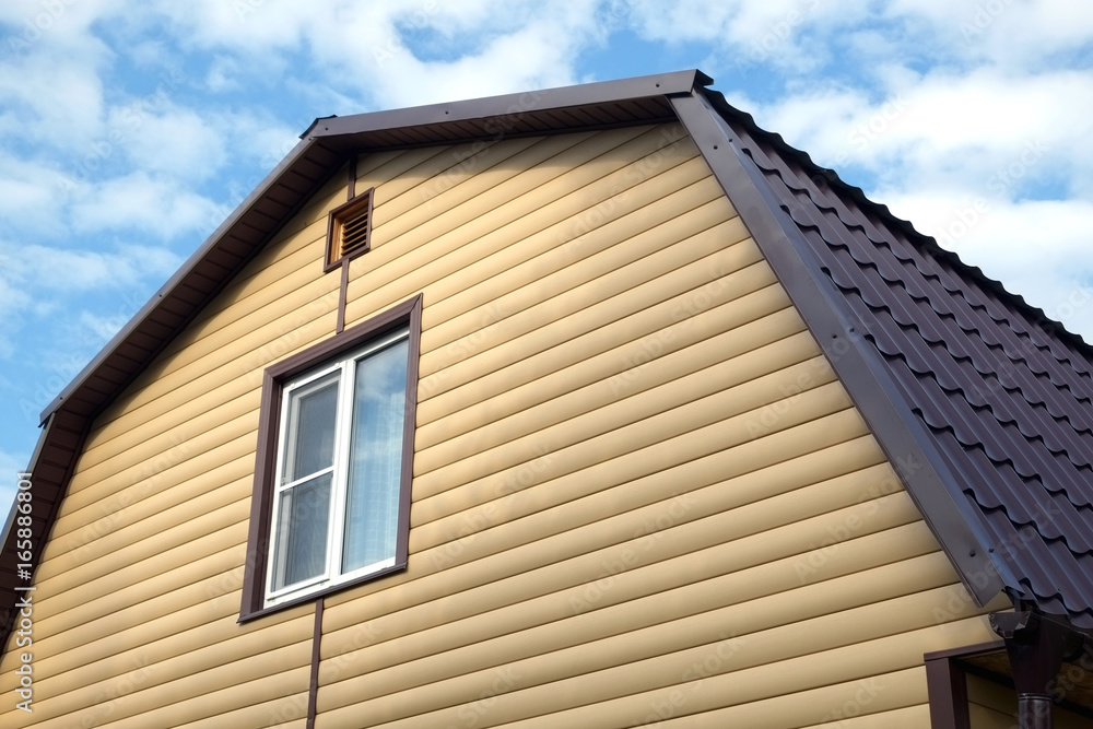 Top part of rural house wall covered with yellow siding and brown metal roof on blue sky with clouds on sunny day front view closeup
