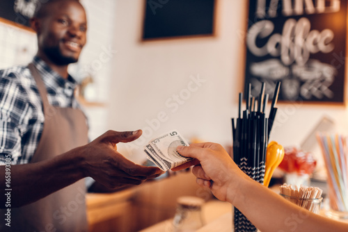 handsome smiling african american barista taking cash payment on bar counter in cafe photo