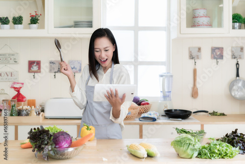Cheerful Asian young woman is cooking in the kitchen with joy. She is standing and holding digital tablet of recipe. Asian woman is touching a wood spoon to her face and thinking.