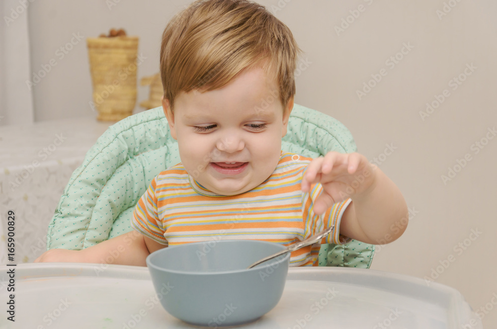 little blond boy in a striped T-shirt eats porridge with a spoon from a gray plate little blond boy in a striped T-shirt eats porridge with a spoon from a gray plate