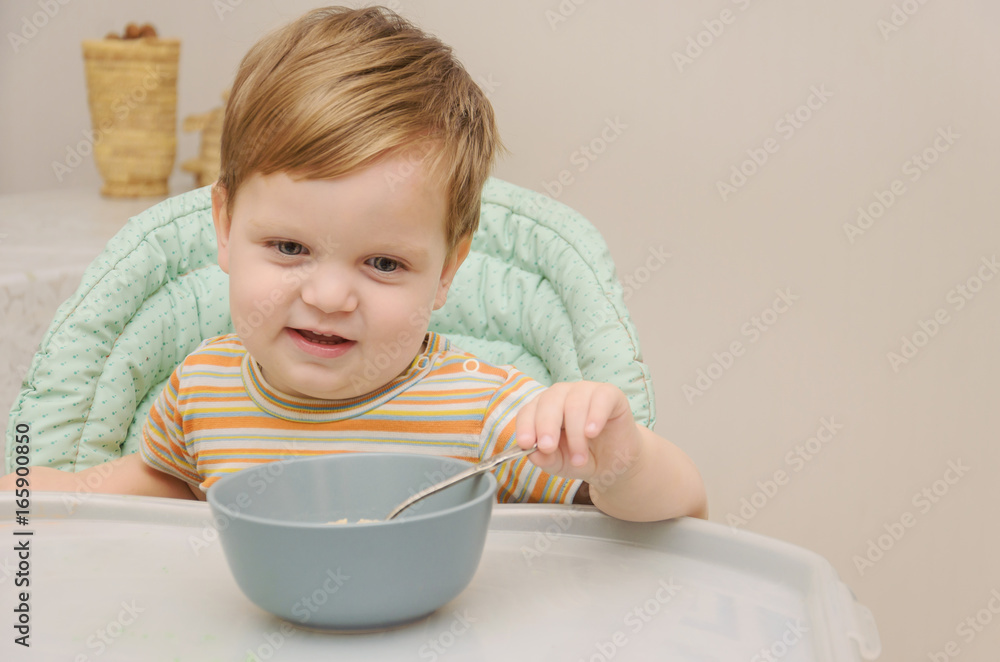 little blond boy in a striped T-shirt eats porridge with a spoon from a gray plate little blond boy in a striped T-shirt eats porridge with a spoon from a gray plate