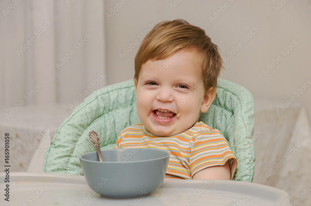little blond boy in a striped T-shirt eats porridge with a spoon from a gray plate little blond boy in a striped T-shirt eats porridge with a spoon from a gray plate