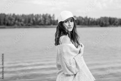 A beautiful well-groomed secured young woman in a casual style of clothes, a knitted cape, a white dress and a cap rests by the water for the weekend and feeling freedom © T.Den_Team