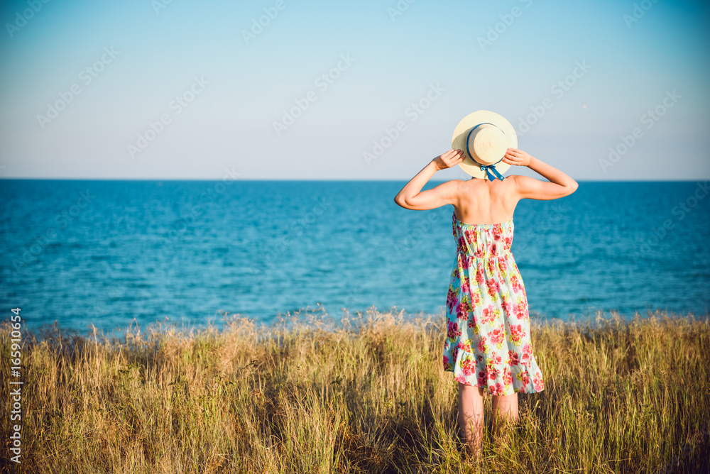 Summer portrait of young pretty woman in a straw hat and dress standing backwards in the grass and looking to the sea. Girl enjoy nature sea paradise and fresh air at wonderful summer vacation