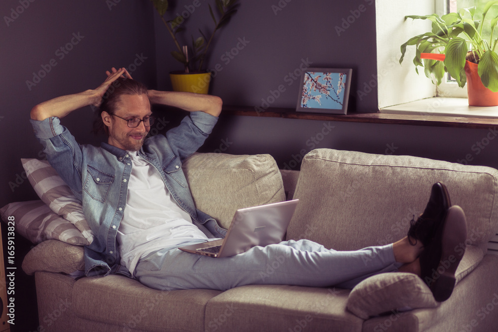 Happy young freelancer lying at couch with laptop. Man in shoes at sofa, watching video. Toned image.