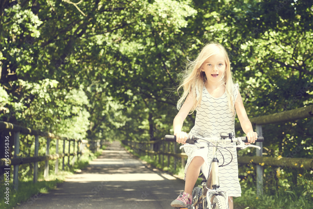 Fototapeta premium Child blond girl riding a bicycle on a bike path in summer