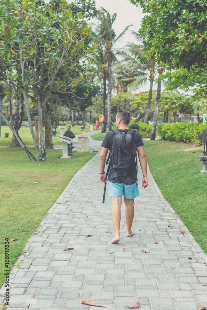 Rear view of man with backpack walking in the beach park of Nusa Dua, Bali island, Indonesia.