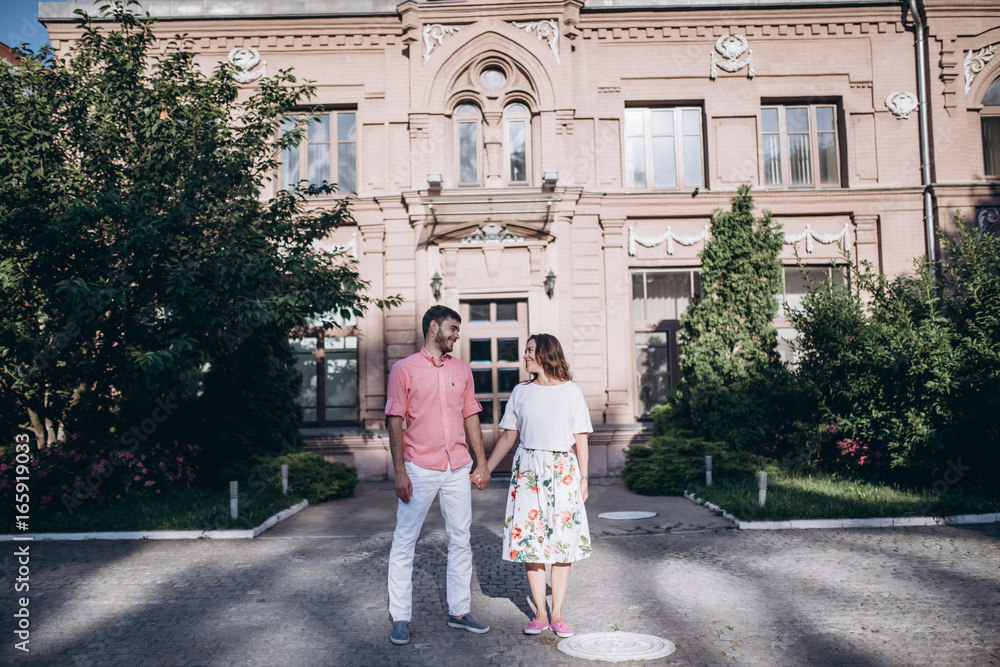 Fototapeta premium Couple in love stand in the old city. Old building and green trees on the background. Couple holding hands and looking at each other.