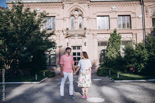 Couple in love stand in the old city. Old building and green trees on the background. Couple holding hands and looking at each other.