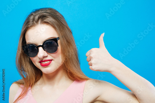 Cute Brunette Wearing Pink Dress  Sunglasses and Sport Shoes is Showing Stop by Hands on Blue Background in Studio.
