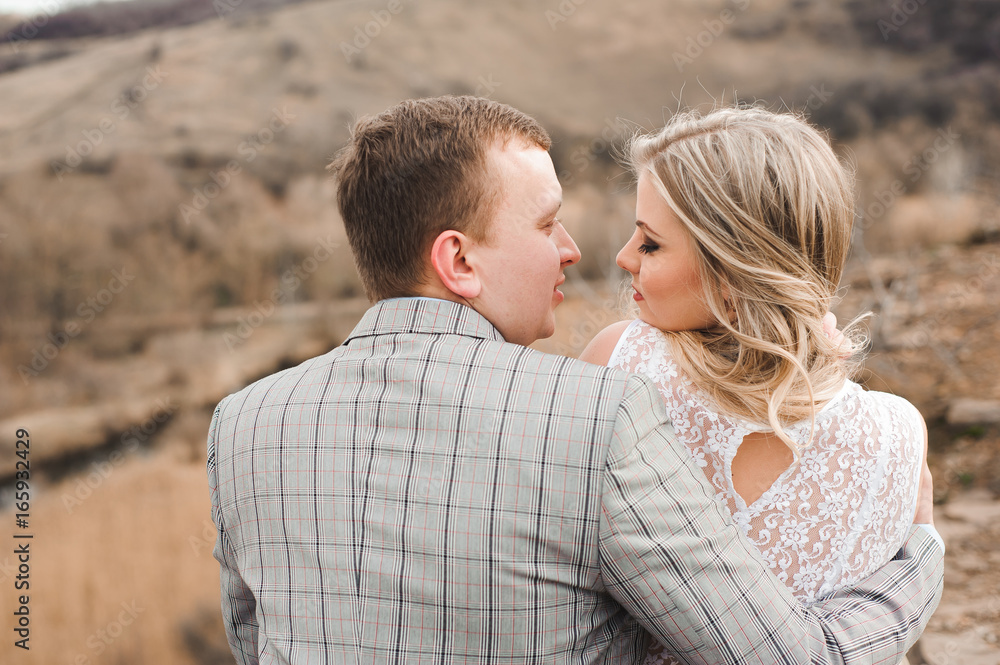Love, romance and people concept - happy young couple hugging sitting on the edge of a cliff outdoors Love, romance and people concept - happy young couple hugging sitting on the edge of a cliff outdoors