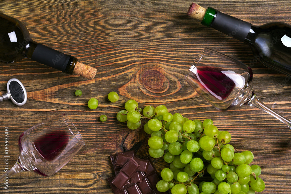 Obraz premium Red and white wine bottles, grape, chocolate and glasses over wooden table. Top view with copy space