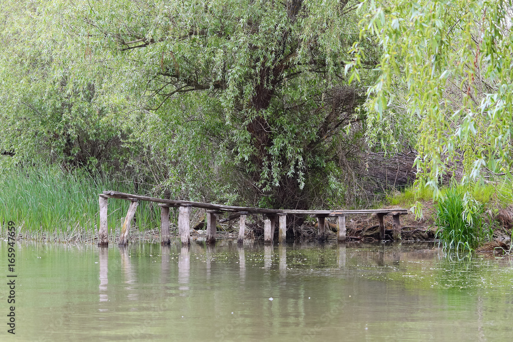 Detail of an old, unpainted wooden pier with calm and still water and green trees in background. Jetty is empty and the wood is weathered and gnarled. Poles are cracked. Detail of an old, unpainted wooden pier with calm and still water and green trees in background. Jetty is empty and the wood is weathered and gnarled. Poles are cracked.
