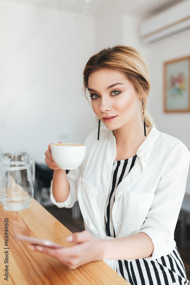 Portrait of young beautiful girl looking in camera with cup of coffee and mobile phone in hands at cafe. Pretty lady with blond hair sitting at the counter and drinking coffee in coffee shop Portrait of young beautiful girl looking in camera with cup of coffee and mobile phone in hands at cafe. Pretty lady with blond hair sitting at the counter and drinking coffee in coffee shop