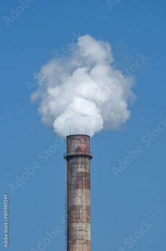 Obsolete vintage industrial chimney with white smoke on blue sky background
