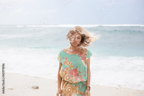 Horizontal portrait of blonde girl in colorful dress with hair floating in the air is standing near the ocean and looking down © Dana Keli