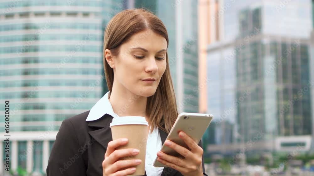 Young successful businesswoman in suit using smart phone in city downtown, professional female employer browsing chatting reading news drinking coffee. Business district skyscrapers in background Young successful businesswoman in suit using smart phone in city downtown, professional female employer browsing chatting reading news drinking coffee. Business district skyscrapers in background