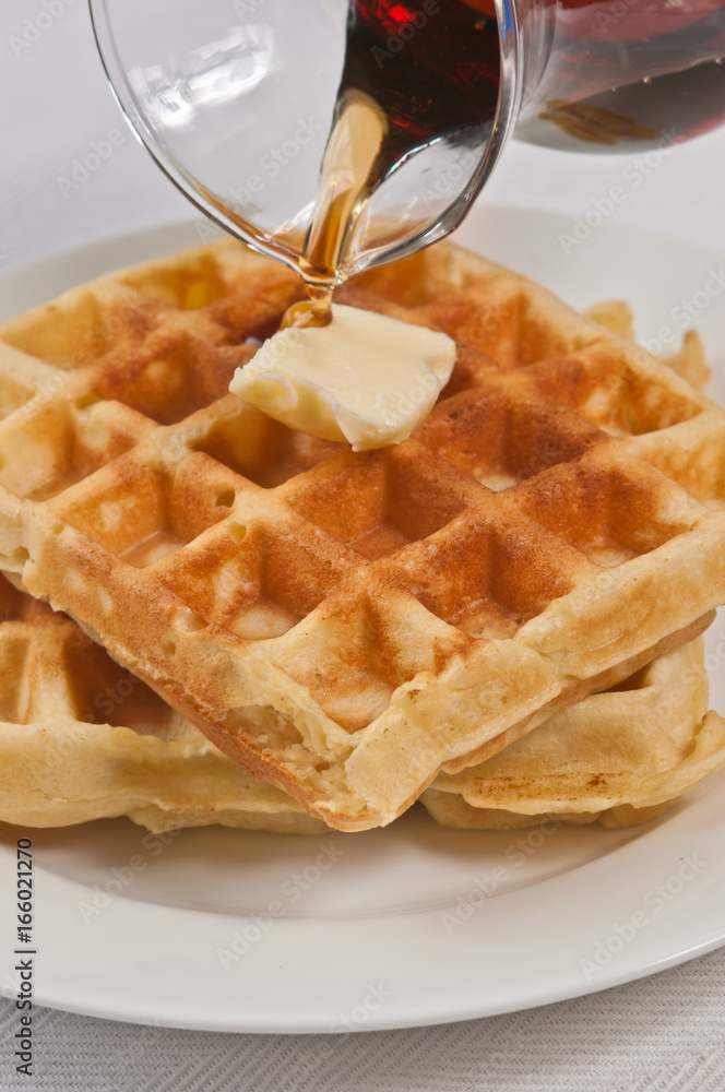 Stack of baked, golden buttermilk waffles on a round, white plate with a pat of butter on top and warm, maple syrup pouring from a clear, glass pitcher Stack of baked, golden buttermilk waffles on a round, white plate with a pat of butter on top and warm, maple syrup pouring from a clear, glass pitcher
