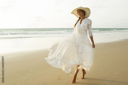 Fotografie Woman wearing beautiful white dress is walking on the beach during sunset