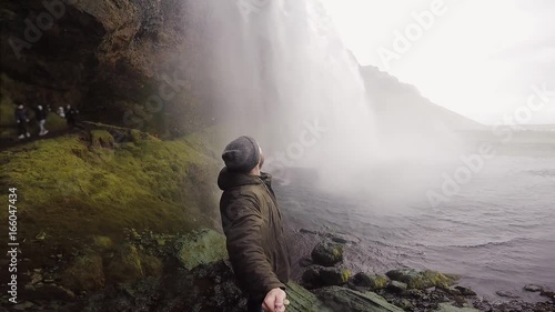 Young tourist man with selfie stick filming on gopro camera powerful waterfall Seljalandsfoss in Iceland in overcast day