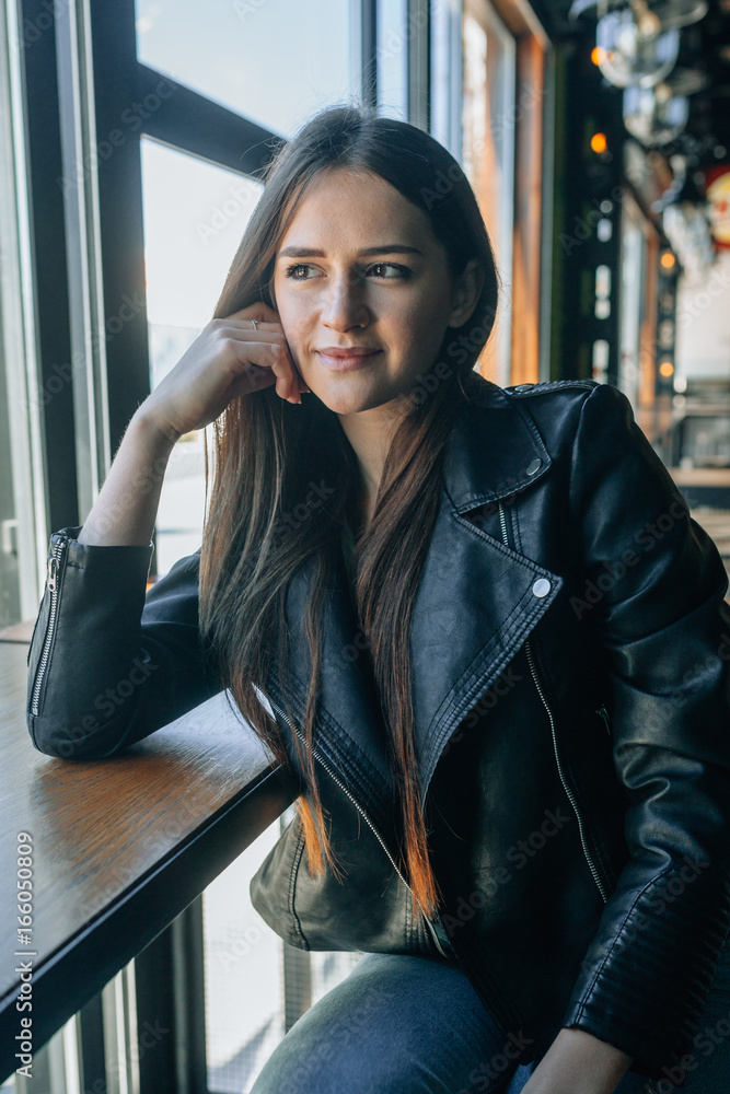 The attractive girl in a black leather jacket and tremendous dark hair sits  in cafe. She looks out of the window the dreaming look and lovely smiles.  Stock Photo | Adobe Stock