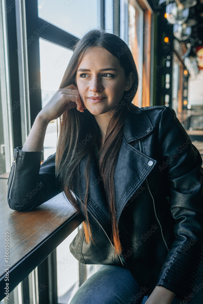 The attractive girl in a black leather jacket and tremendous dark hair sits in cafe. She looks out of the window the dreaming look and lovely smiles. Stock Photo Adobe Stock