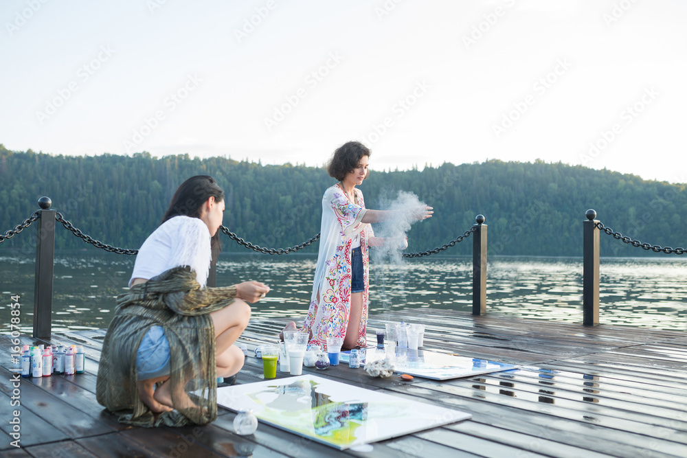 Fototapeta premium Two young happy fashion women painting a picture on an easel on a warm sunny day at nature. Art therapy, relax and hobby concept