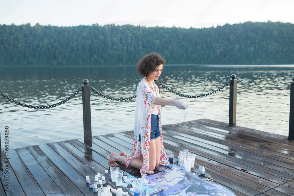 Young beautiful artist woman is painting with acrylic paint on open air session. Art therapy on wooden pier at nature lake bay