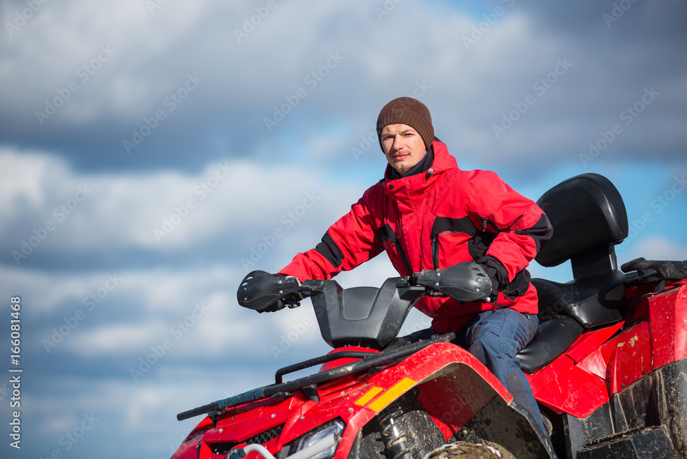 Close-up portrait of man in a red winter clothes riding on red quad bike in the mountains top. Blue sky with clouds on the background