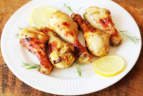 Grilled chicken legs with mustard on wooden table served on white plate with rosemary and lemon. BBQ dinner background