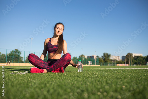 Young pretty sportswoman in sportswear sitting on stadium green grass outdoors with bottle of water. Healthy lifestyle concept. Summer sport activity.