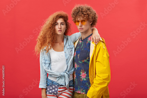 Style and fashion. Attractive confident young male and female models posing at blank studio wall, dressed in trendy clothes, looking at camera with serious facial expressions; girl hugging guy